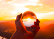 una mano tiene una sfera trasparente attraverso la quali si vede il tramonto all'orizzonte